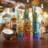 set of 16oz multicolor Tiki glasses siting next to coconut and full of beverage.