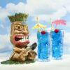 two tiki glasses with an electric blue beverage next to a carved tiki statue.