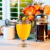 a glass of orange juice in a 10 oz glass goblet on a table with a cup of coffee and a press.