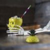 A skeleton arm holding a yellow pig skull tiki glass on a table. The glass has a swizzle straw in it and is sitting next to the pineapple top.